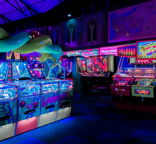 An indoor arcade filled with vibrant gaming machines lit with neon blue and pink lights. The scene includes various arcade games with flashing screens and colorful displays, and a large overhead structure resembling a green snake. The background shows illuminated signs and themed decor, contributing to an immersive entertainment atmosphere.