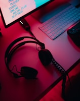 A dimly lit gaming setup with a monitor displaying a game's menu on the screen. A mechanical keyboard sits in front of the monitor. A pair of over-ear headphones and a game controller rest on the desk. The scene is bathed in a red and blue light, creating a moody gaming atmosphere.