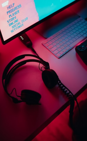 A dimly lit gaming setup with a monitor displaying a game's menu on the screen. A mechanical keyboard sits in front of the monitor. A pair of over-ear headphones and a game controller rest on the desk. The scene is bathed in a red and blue light, creating a moody gaming atmosphere.