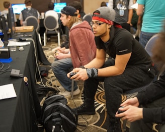 Several people are sitting in a room with gaming consoles and monitors, focusing intently on their screens. The setting appears to be a casual gaming event, with participants holding controllers while playing video games. The room has a casual atmosphere with chairs and tables set up for gaming.