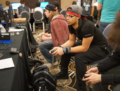 Several people are sitting in a room with gaming consoles and monitors, focusing intently on their screens. The setting appears to be a casual gaming event, with participants holding controllers while playing video games. The room has a casual atmosphere with chairs and tables set up for gaming.