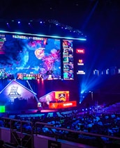 A large esports arena featuring a massive screen displaying a video game, surrounded by sponsor logos and team names. The auditorium is filled with spectators watching the gaming event. Neon blue and purple lights illuminate the stage, creating a vibrant atmosphere.