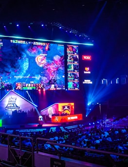 A large esports arena featuring a massive screen displaying a video game, surrounded by sponsor logos and team names. The auditorium is filled with spectators watching the gaming event. Neon blue and purple lights illuminate the stage, creating a vibrant atmosphere.