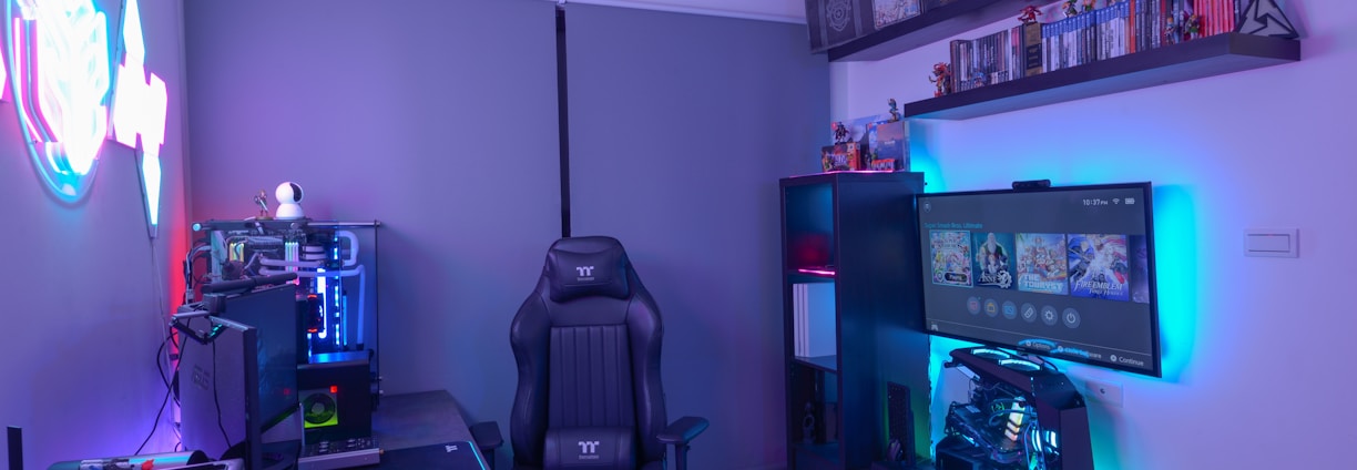A modern gaming setup featuring a sleek gaming chair, dual monitors on a desk, and LED lighting. A gaming console and various decorations are displayed on shelves, creating an organized and vibrant gaming environment.