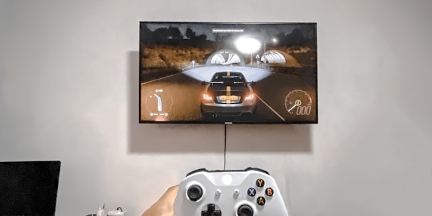 A person is holding a gaming controller in front of a flat-screen television displaying a racing video game. The room has a computer setup on a wooden desk with cables, a desktop computer monitor, and other accessories. The focus is on the interaction between the person and the gaming environment.