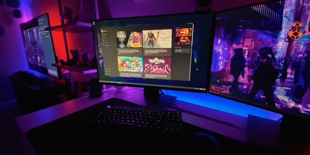 A gaming setup featuring multiple screens with vibrant game-related images illuminated by colorful LED lighting. The environment is dimly lit, highlighting the vivid displays on the monitors. A keyboard is visible on the desk beneath the central monitor.