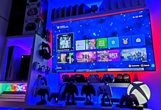 A modern gaming setup featuring a large wall-mounted screen displaying a gaming console interface. The room is decorated with gaming-themed items such as controllers and collectibles. The ambient lighting includes shades of blue and red, creating a vibrant atmosphere.