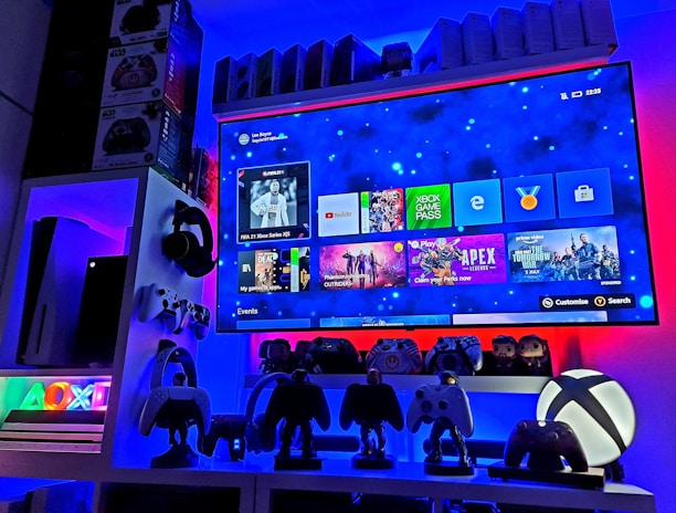 A modern gaming setup featuring a large wall-mounted screen displaying a gaming console interface. The room is decorated with gaming-themed items such as controllers and collectibles. The ambient lighting includes shades of blue and red, creating a vibrant atmosphere.
