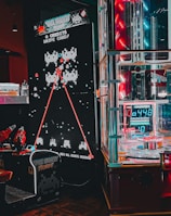 An arcade setting featuring a classic Space Invaders game setup and a modern gaming machine with seats. Bright neon lights illuminate the area, with scores displayed on digital screens. A bright and visually engaging environment full of electronic components.