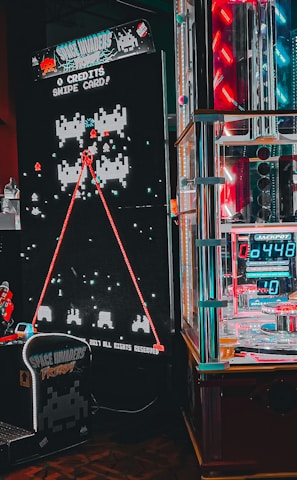 An arcade setting featuring a classic Space Invaders game setup and a modern gaming machine with seats. Bright neon lights illuminate the area, with scores displayed on digital screens. A bright and visually engaging environment full of electronic components.