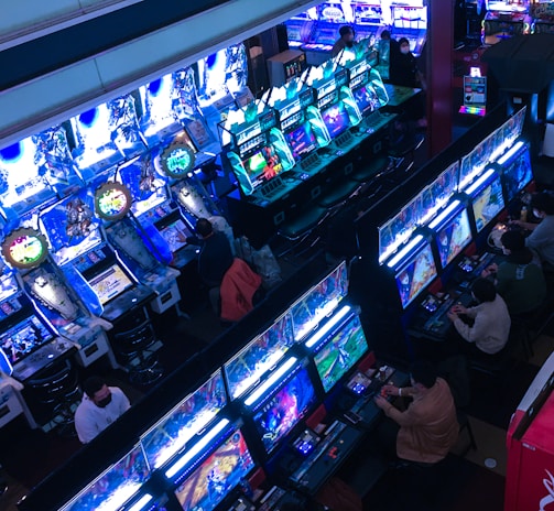 A bustling arcade filled with rows of gaming machines, each with vibrant displays and controls. Several people are seated at these machines, deeply engaged in playing games. Bright fluorescent lights cast a blue and neon glow across the room, enhancing the lively and energetic atmosphere.