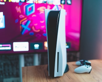 A gaming console is placed on a wooden table with a game controller nearby. In the background, a television screen displays colorful graphics, including geometric shapes and vibrant colors.
