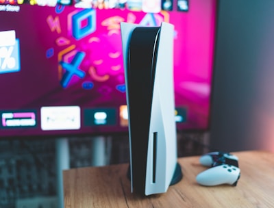 A gaming console is placed on a wooden table with a game controller nearby. In the background, a television screen displays colorful graphics, including geometric shapes and vibrant colors.