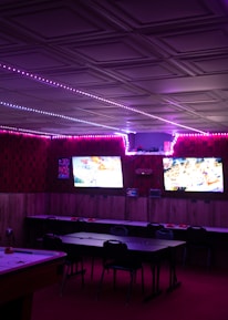 A dimly lit room with vibrant purple LED strip lights on the ceiling, featuring multiple gaming stations. There are two large screens showing video games, along with various arcade machines, including a Nintendo arcade cabinet. An air hockey table is positioned in the foreground.