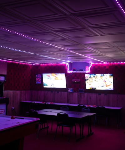 A dimly lit room with vibrant purple LED strip lights on the ceiling, featuring multiple gaming stations. There are two large screens showing video games, along with various arcade machines, including a Nintendo arcade cabinet. An air hockey table is positioned in the foreground.