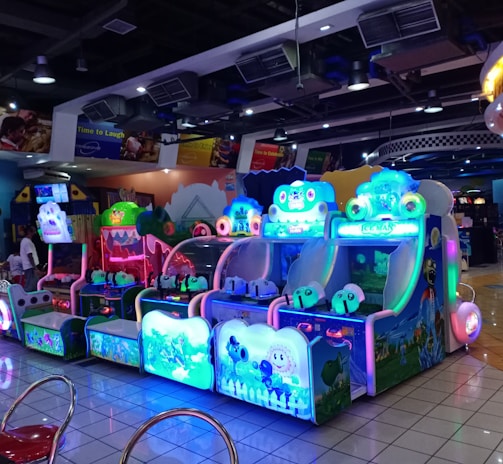 A brightly lit arcade filled with colorful gaming machines sits under ceiling lights. Neon lights illuminate various game stations, creating a playful and vibrant environment. A few red chairs are scattered around the reflective, tiled floor, and a person is observing one of the machines. The atmosphere is lively and cheerful, featuring games with cartoonish designs and engaging themes.