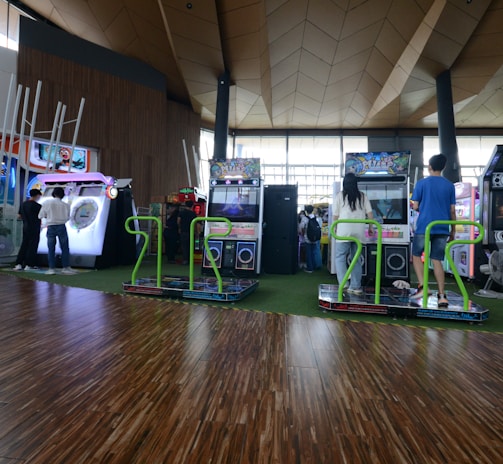 A vibrant arcade setting with several gaming machines lined up, including dance and rhythm game setups. People are engaged with the games, absorbed in their activities. The flooring is wooden, and the ceiling has a geometric design. Natural light filters through large windows, creating a lively atmosphere.