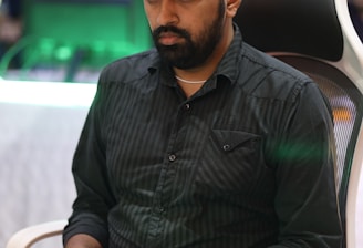 A person is seated in a chair, focused intently on something, with hands positioned on a gaming console or arcade controller. The setup appears to be indoors, with visible screens and lighting in the background, suggesting an electronic gaming environment.