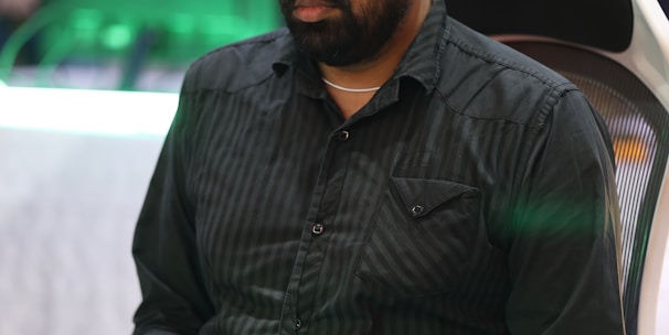 A person is seated in a chair, focused intently on something, with hands positioned on a gaming console or arcade controller. The setup appears to be indoors, with visible screens and lighting in the background, suggesting an electronic gaming environment.